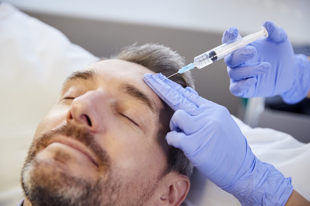 Female Beautician Giving Mature Male Patient Botox Injection In Forehead